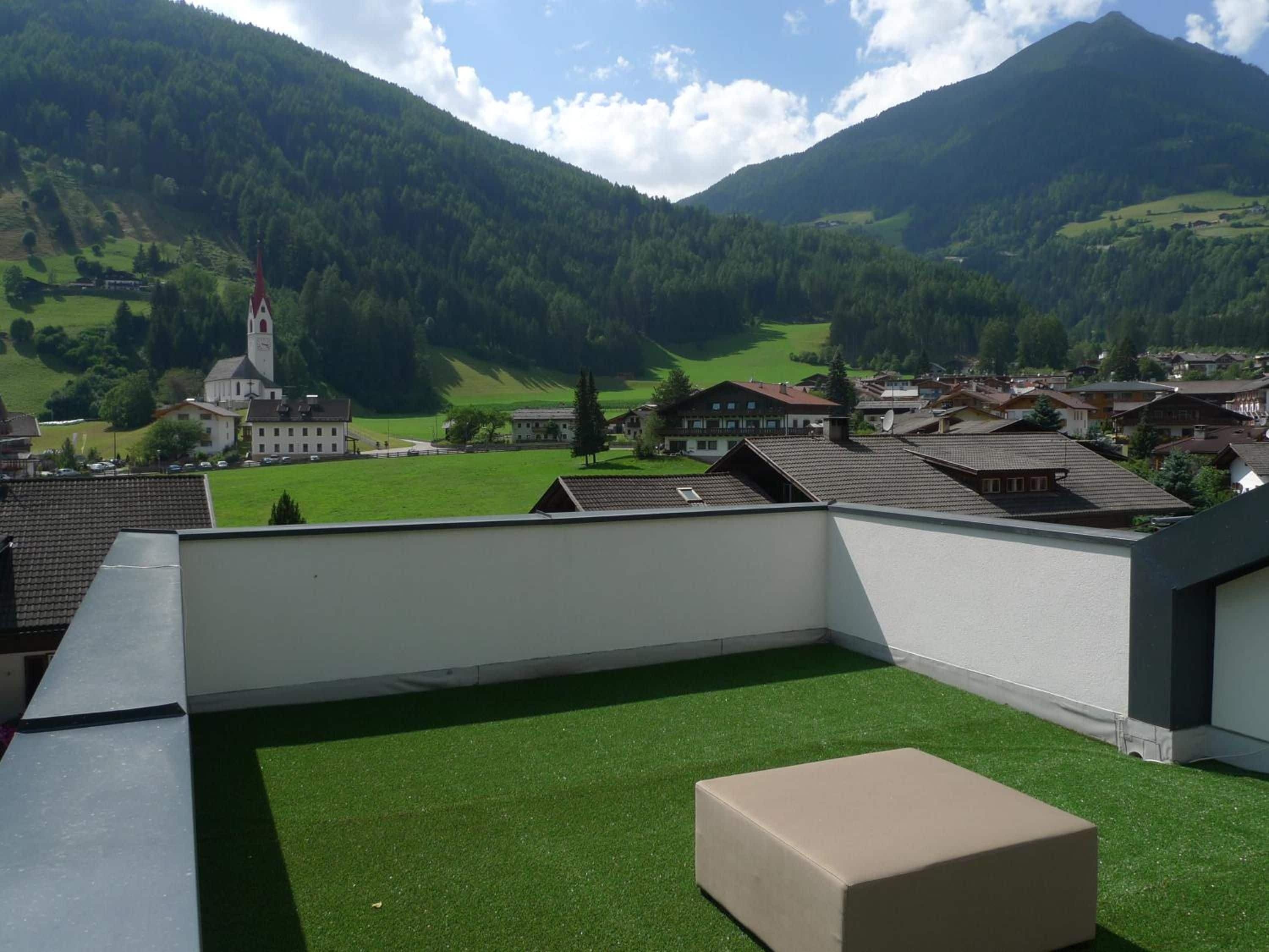 Una terrazza sul tetto con prato sintetico, un poggiapiedi beige e vista su un villaggio con una chiesa e montagne sullo sfondo.