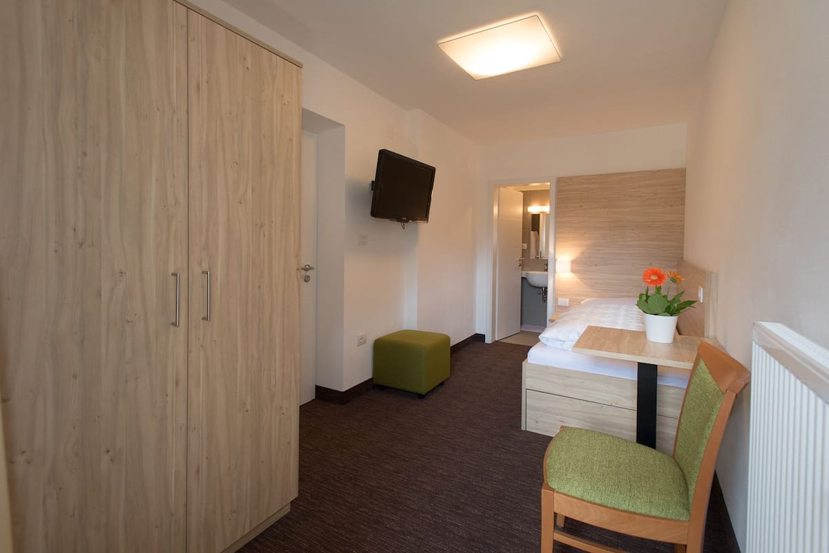 Habitación de hotel con un armario de madera, un otoman verde, una cama con cabecera blanca, una mesita con planta en maceta y un televisor de pantalla plana montado en la pared.