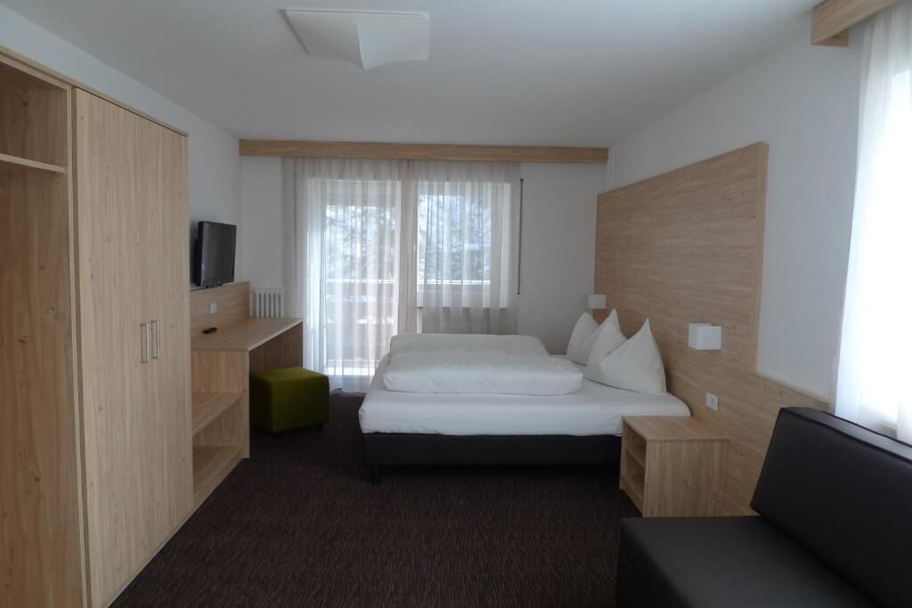 A hotel room with a bed, a TV, a wooden wardrobe, a green ottoman, and a window with curtains.