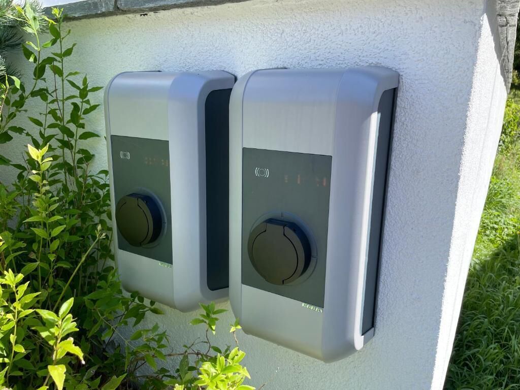 Deux interrupteurs extérieurs, fixés au mur, probablement destinés à contrôler une porte de garage ou un mécanisme similaire, sur un mur de couleur claire, avec de la verdure tout autour.