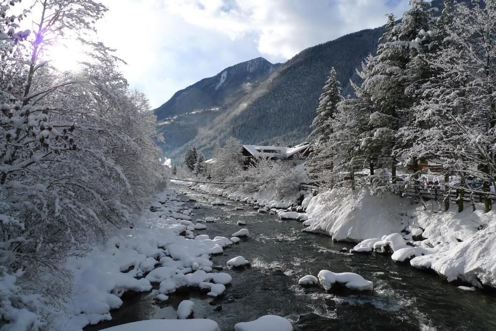 A snow-covered river with a wooden bridge, surrounded by snow-laden trees and mountains in the background.