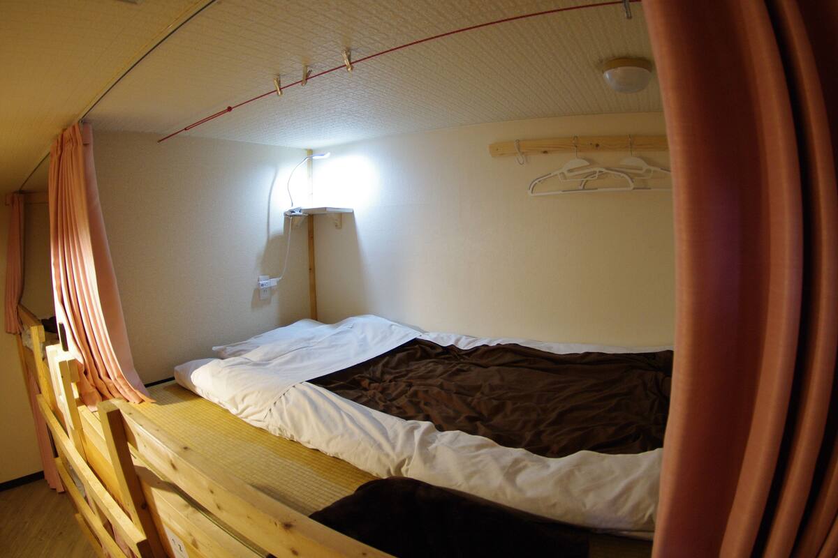 A single bed with a wooden frame, a white mattress, and a brown blanket. There is a wooden shelf above the bed with a hanging light fixture.