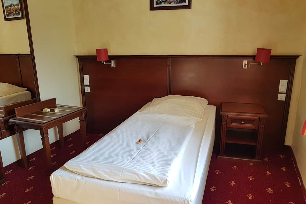 A single bed with a wooden headboard, a small bedside table, a chair, and a red carpeted floor.