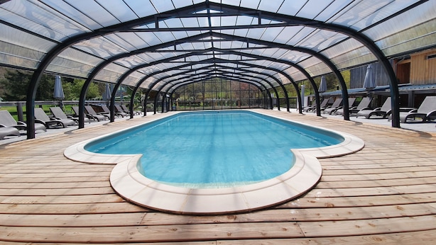 A covered swimming pool with a wooden deck and lounge chairs. - Camping les Vernières (La Bourboule)