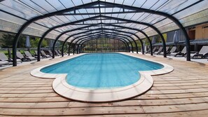 A covered swimming pool with a wooden deck and lounge chairs. - Camping les Vernières (La Bourboule)