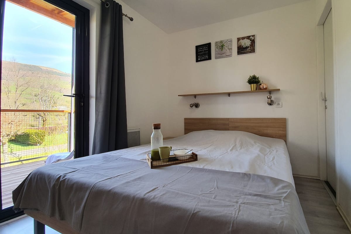 Un dormitorio con cama, una ventana con cortinas, un estante pequeño con adornos y vistas al exterior.
