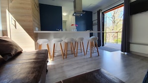 A modern living room with a brown sofa, a dining area with white chairs, and a kitchen counter. - Camping les Vernières (La Bourboule)