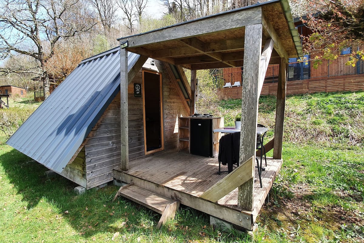 Una pequeña cabaña de madera con techo inclinado, un porche y una mesa con una silla.