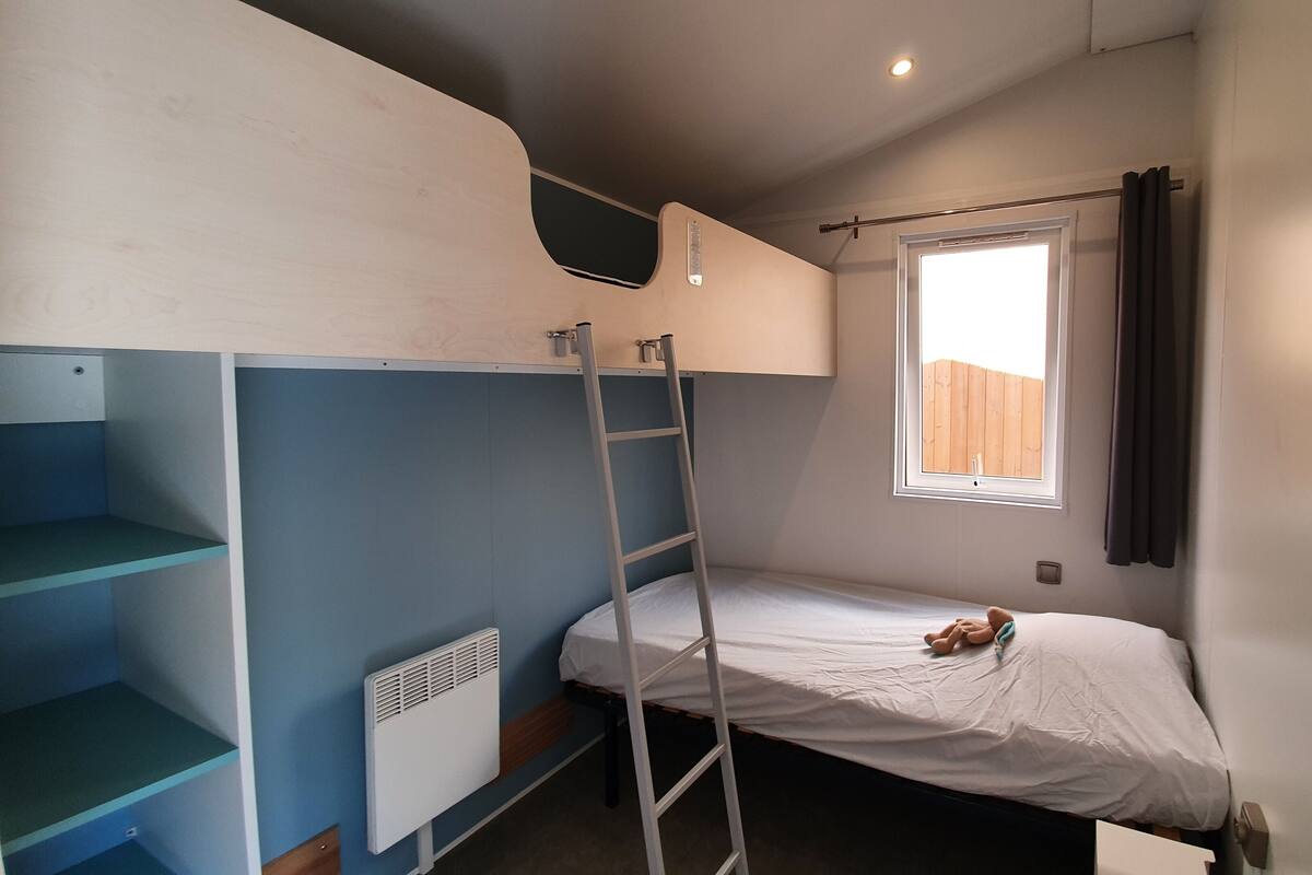 Una habitación pequeña con cama individual y literas a la izquierda, una escalera, una ventana con cortinas y un radiador.