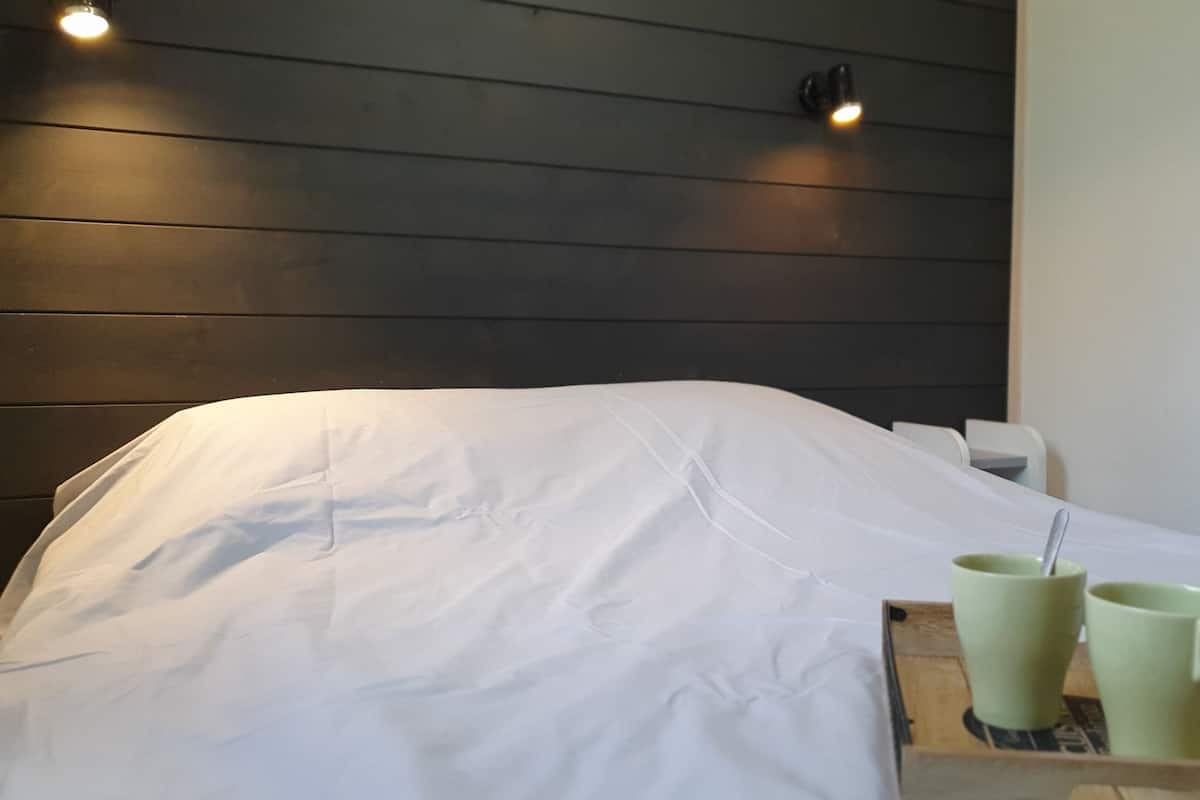 Una cama bien tendida con ropa de cama blanca, una bandeja con dos tazas verdes y una pequeña silla blanca.