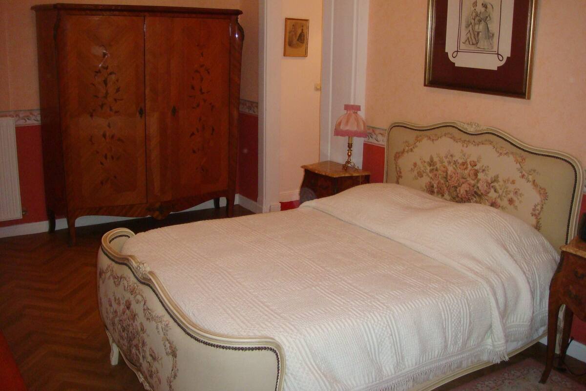 A bedroom with a large wooden wardrobe, a bed with a floral headboard, and a small bedside table with a lamp.
