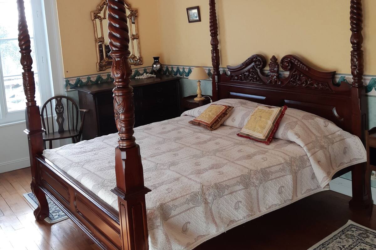 A four-poster bed with a quilted bedspread and two decorative pillows, a wooden nightstand with a lamp, and a chair in a room with wooden flooring and a window.