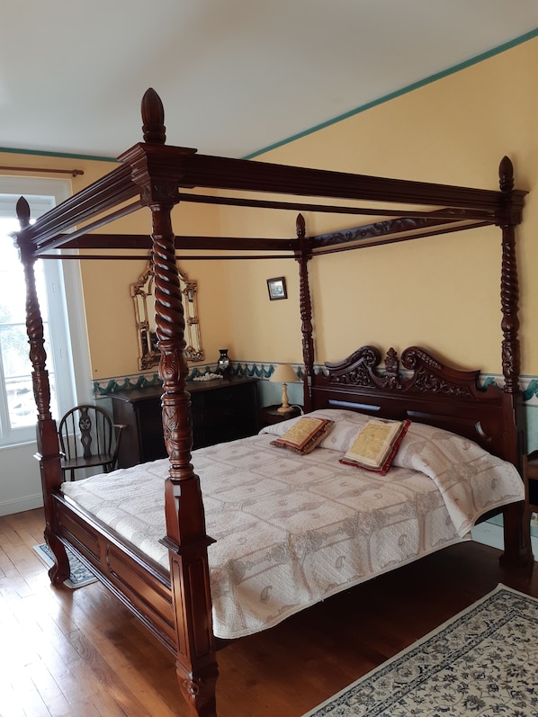 A four-poster bed with a quilted bedspread and two decorative pillows, a wooden nightstand with a lamp, and a chair in a room with wooden flooring and a window.
