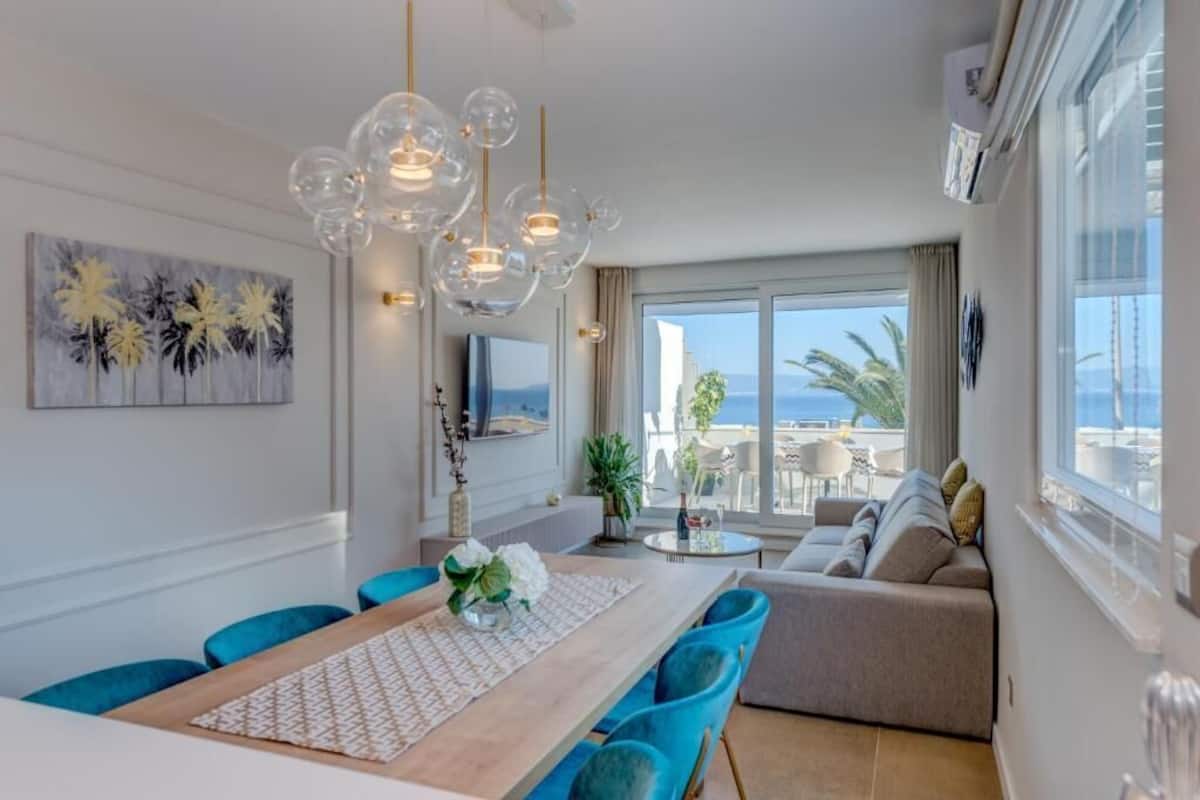 Ein modernes Wohnzimmer mit großem Fenster, einem Essbereich mit Holztisch und blauen Stühlen sowie einer Couch mit Kissen.
