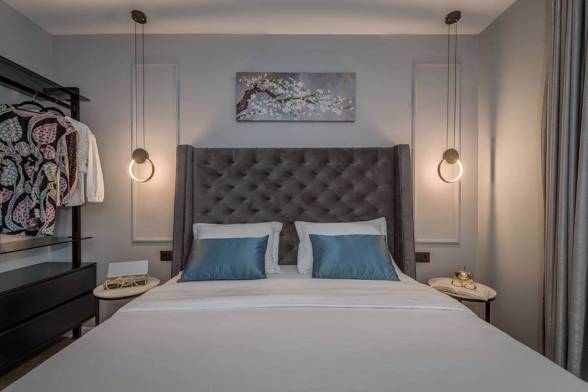 Ein ordentlich eingerichtetes Schlafzimmer mit einem gepolsterten Kopfteil, einem großen Bett mit zwei Kissen, Nachttischen mit Lampen und einem an der Wand hängenden Kunstwerk.