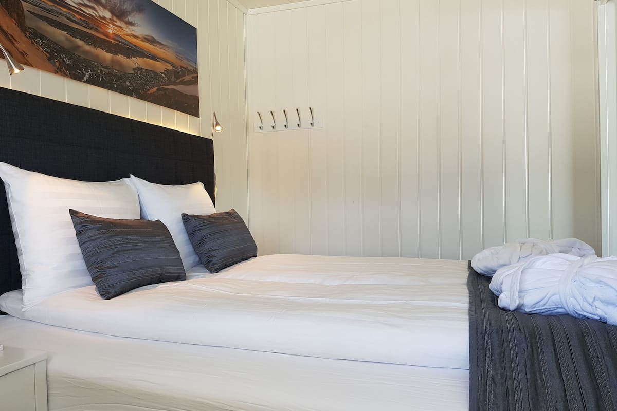 A neatly made bed with white linens and dark pillows, a headboard with a cityscape painting, a bedside table with a lamp, and a wooden bench with a towel.