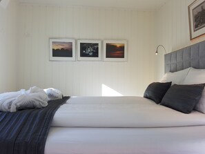 Ein ordentlich bezogenes Bett mit grauem Kopfteil, weißer Bettwäsche und dunklen Kissen. Darüber an der Wand hängen drei gerahmte Bilder.