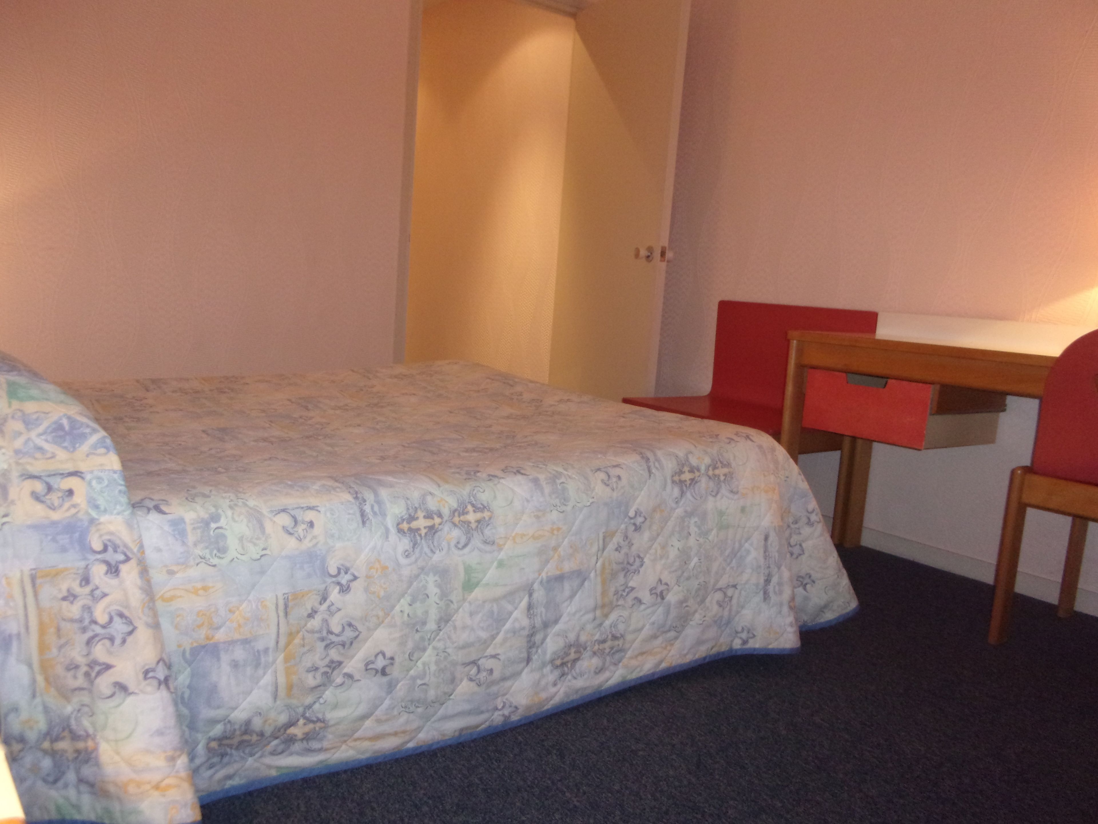 Ein kleines Zimmer mit einem Einzelbett, einer karierten Bettdecke, einem Holztisch und einem roten Stuhl.