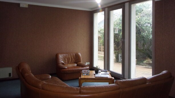 A living room with a brown leather sofa, a wooden coffee table, and a brown leather armchair. There is a large window with a view of greenery outside. - Jackotel Orleans (Orléans)