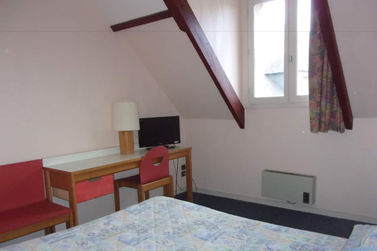 Quarto com cama, escrivaninha, cadeira, televisão e janela com cortinas.