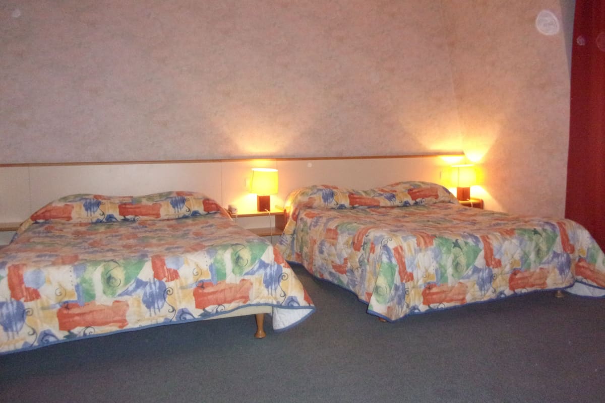 Quarto de hotel com duas camas, cada uma com colcha estampada, duas luminárias de cabeceira e cortina vermelha.