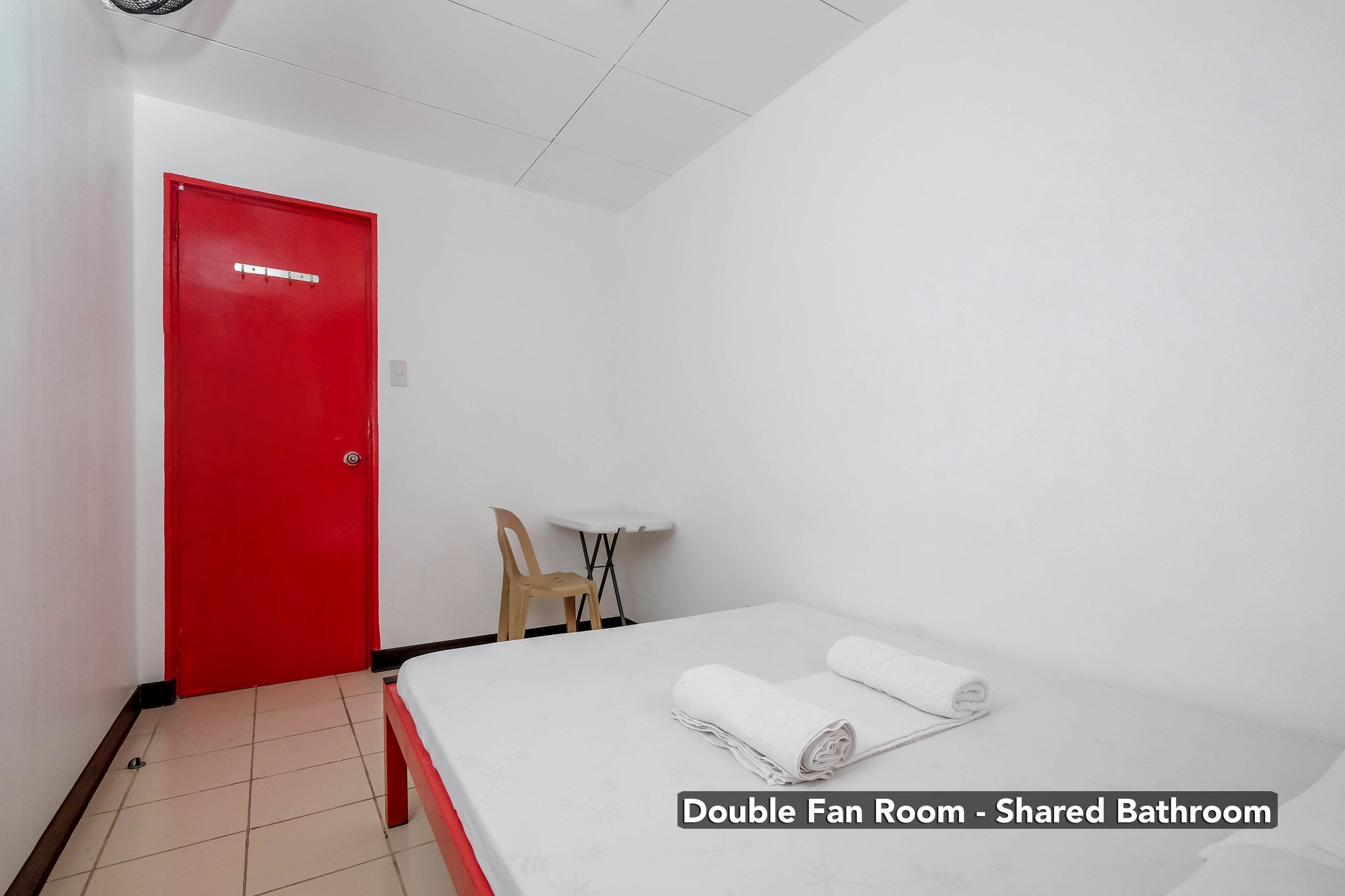 Habitación doble con ventiladores, puerta roja, cama con ropa blanca, una mesita y una silla.