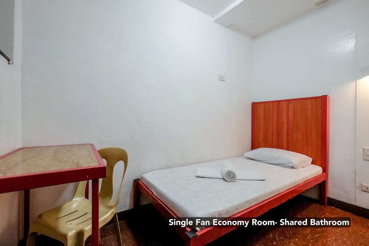 A single fan economy room with a shared bathroom, featuring a bed with a wooden headboard, a small table, and a chair.