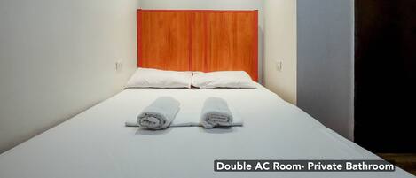 A double AC room with a private bathroom, featuring a bed with white linens and two rolled towels, a wooden headboard, and a light-colored wall.