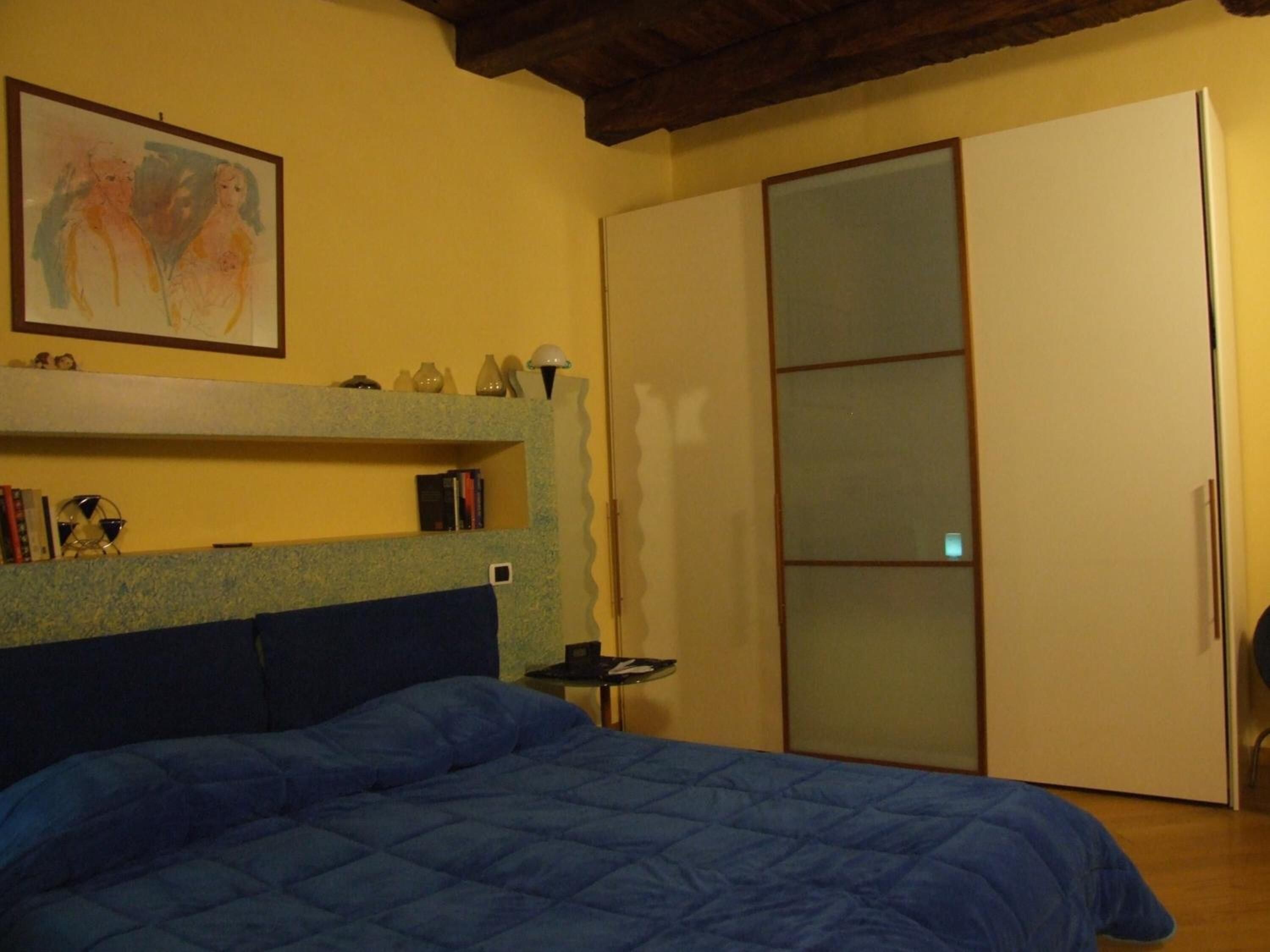 A bedroom with a bed, a bookshelf, a painting, and a wardrobe.