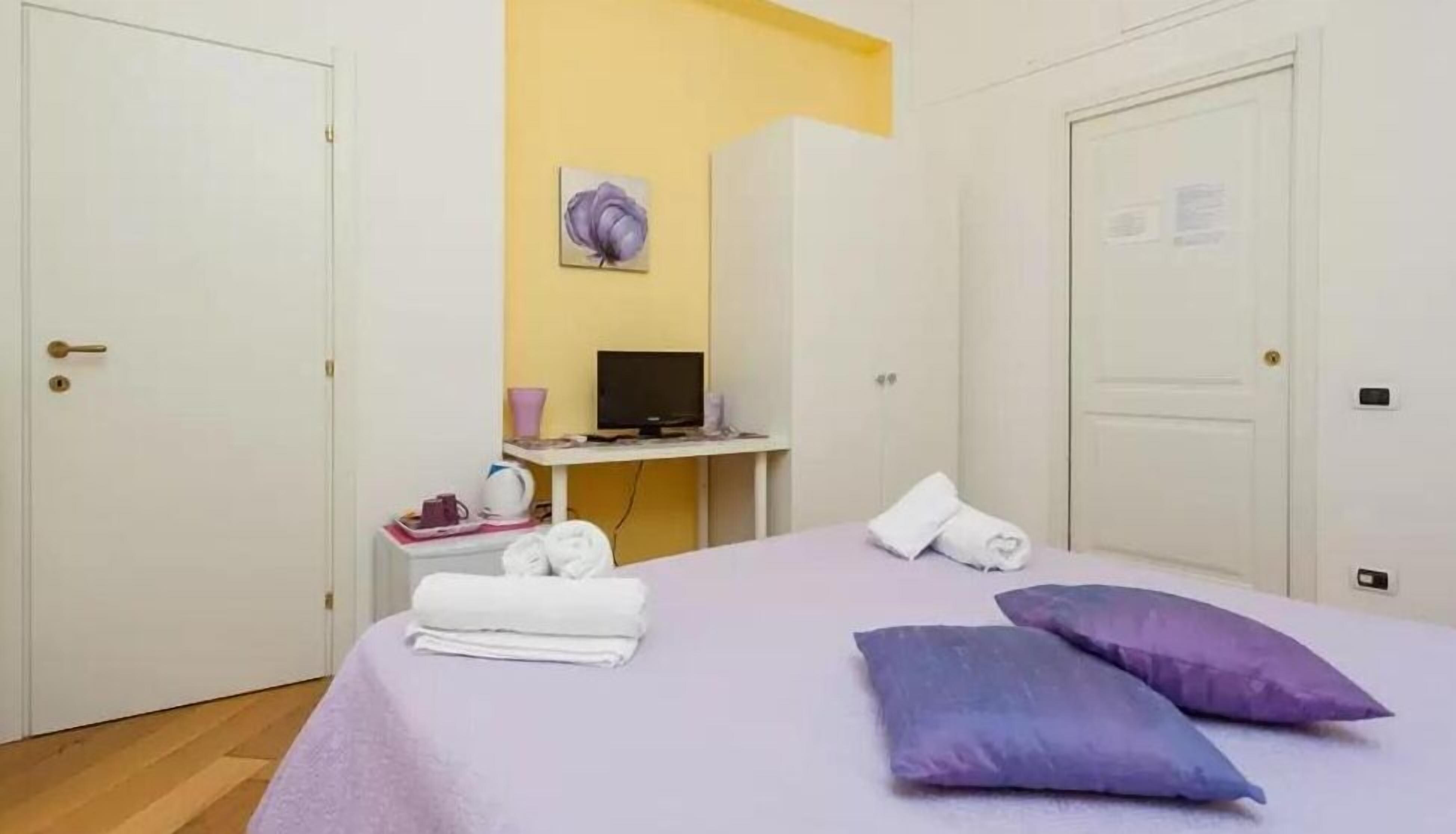 A hotel room with a bed, a desk with a computer, a chair, and a small table with a lamp.