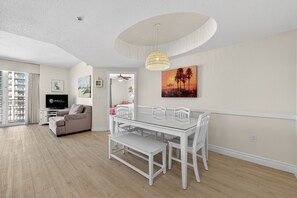 Een moderne woonkamer met een glazen eettafel, witte stoelen, een beige bank en een groot raam.