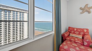 A room with a large window overlooking a beach and a high-rise building. There is a red chair with a patterned cushion and a starfish decoration on the wall.