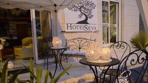 Un área de estar al aire libre con sillas y mesas de mimbre, iluminada por faroles decorativos. Se ve un cartel de 'HotelsSilva' al fondo.