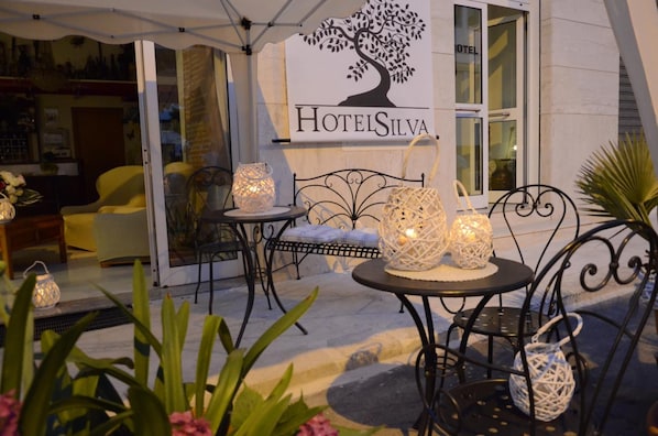 An outdoor seating area with wicker chairs and tables, illuminated by decorative lanterns. A sign for 'HotelsSilva' is visible in the background.