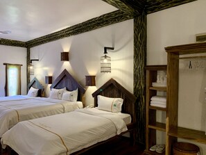 A hotel room with two beds, wooden headboards, a wardrobe, and a shelf with folded towels.