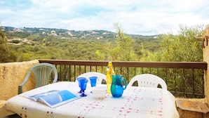 Une table dressée avec de la vaisselle bleue et jaune, un pichet blanc et un livre, le tout placé sur un balcon donnant sur un paysage vallonné.