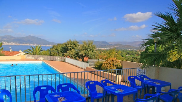 Un espace piscine avec des chaises et des tables bleues, entouré d’une clôture métallique, offrant une vue panoramique sur un paysage magnifique avec des montagnes et une étendue d’eau.