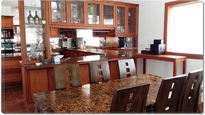 A kitchen with wooden cabinets, a granite countertop, and a dining area with chairs. - Wongsuwan Pool Villa (Hua Hin)