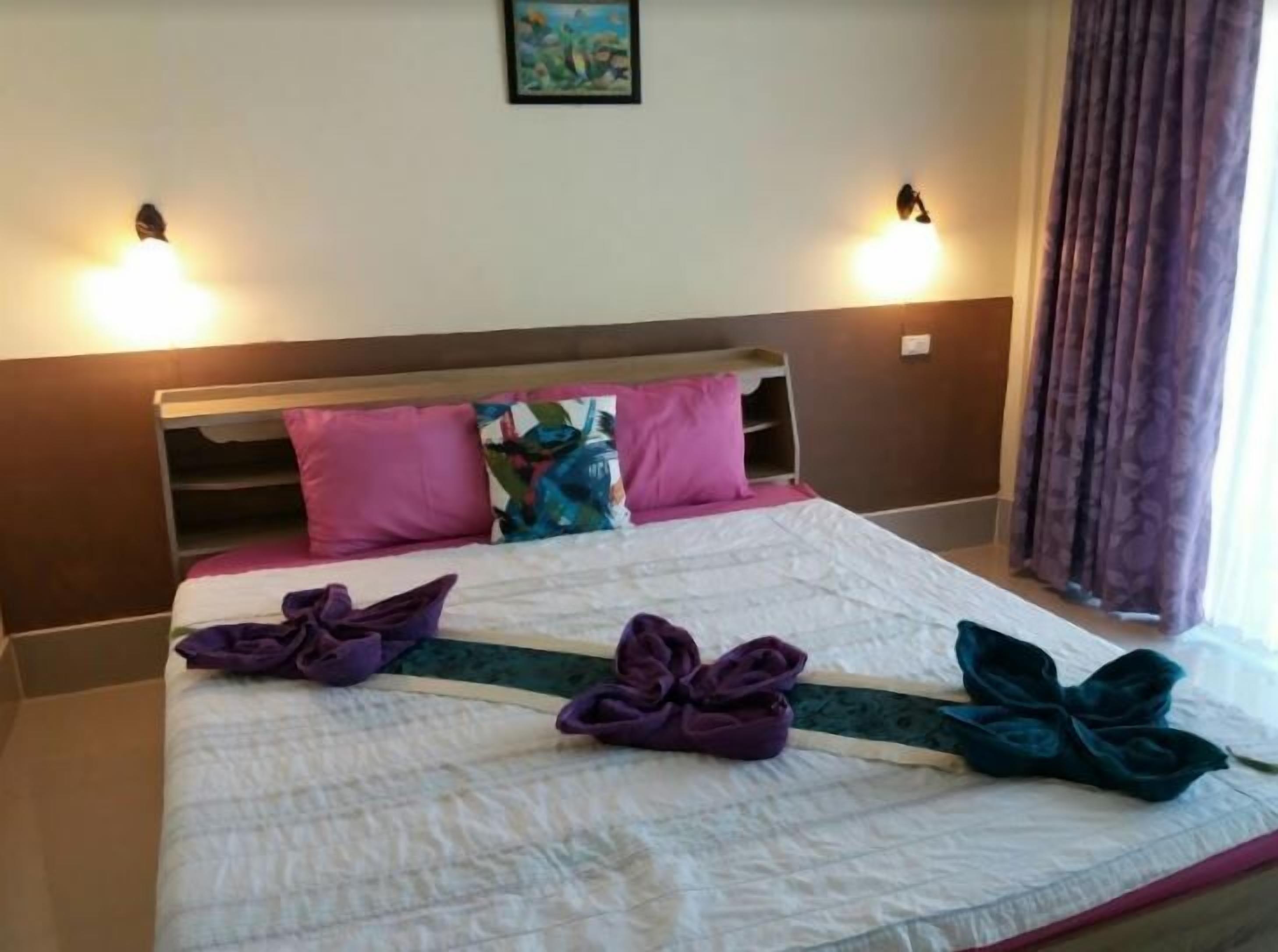 A hotel room with a bed, two wall-mounted lamps, a painting above the bed, and a window with curtains.