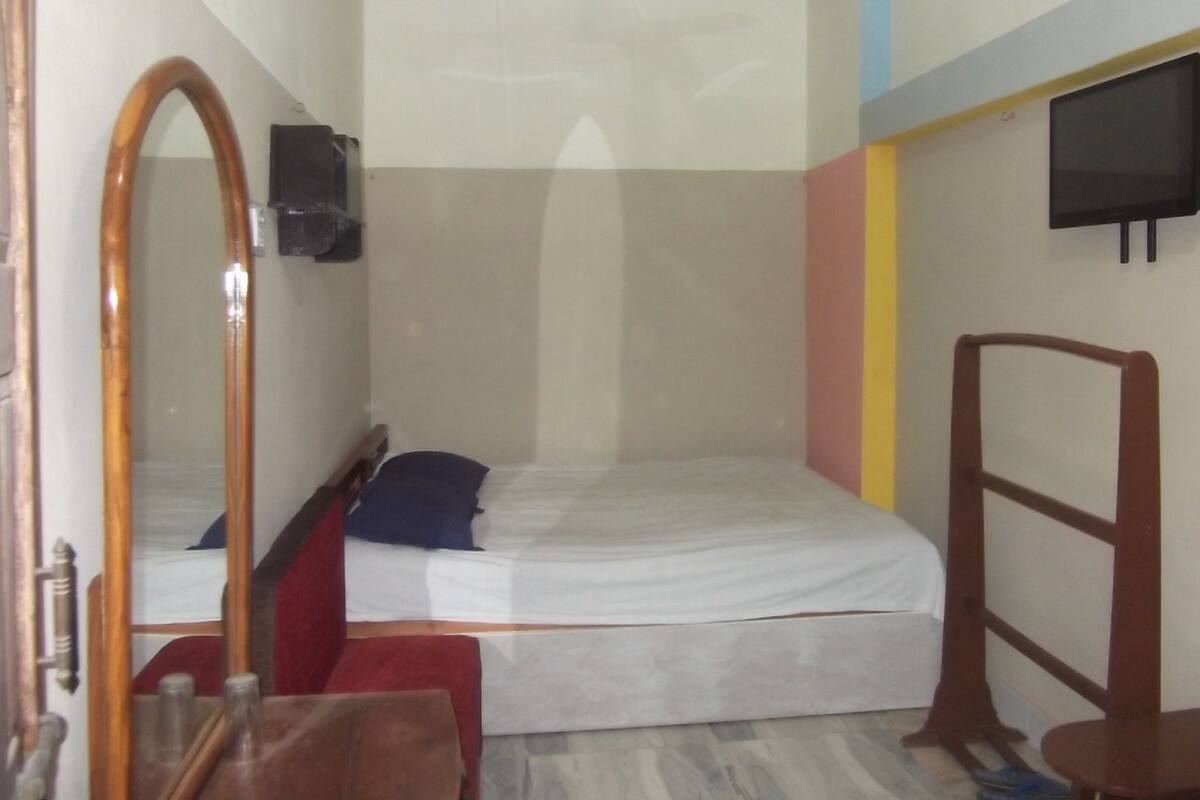 A small, single-bed room with a wooden headboard, a mirror, a television on the wall, and a small table.