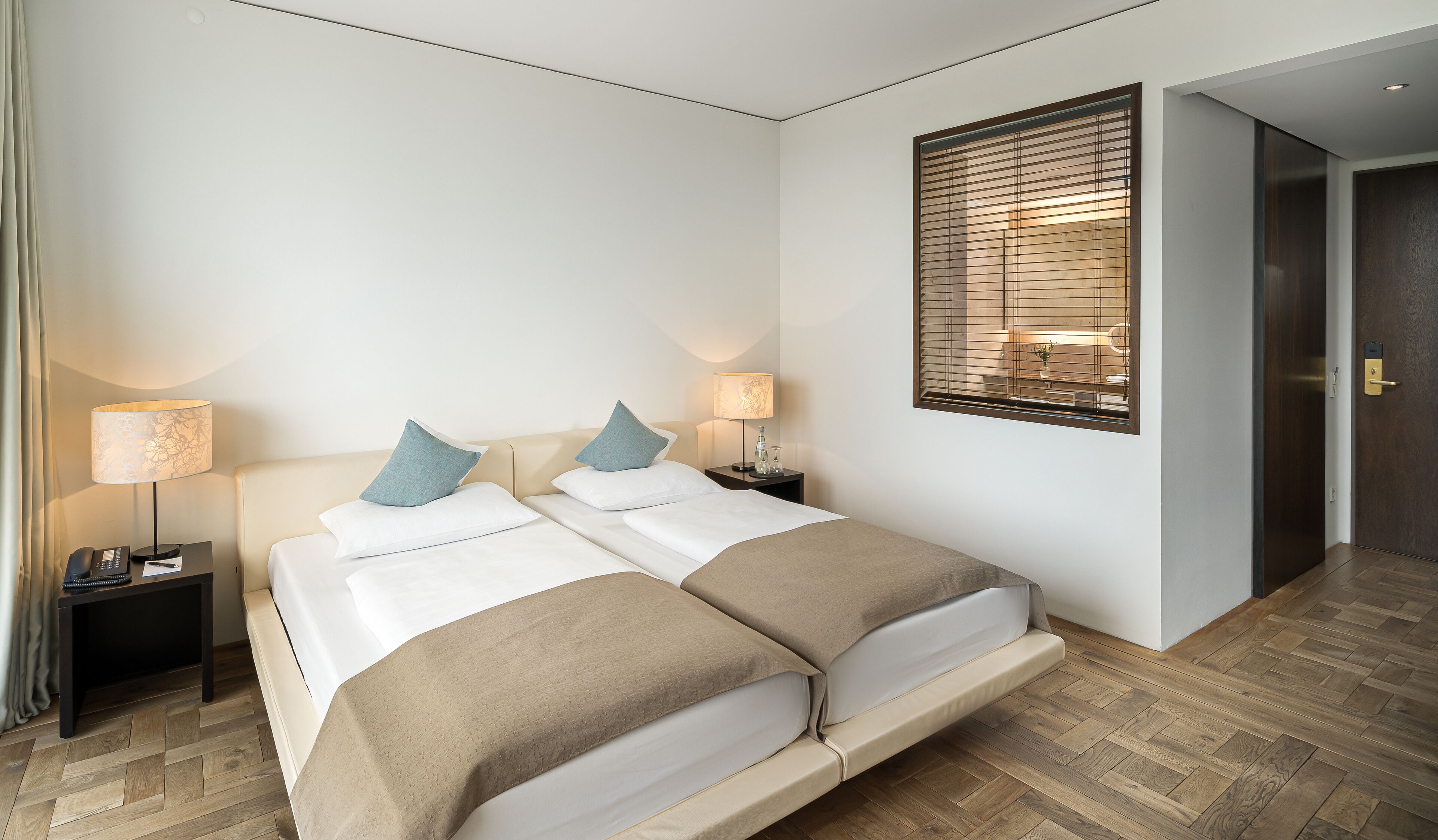 A hotel room with a double bed, two bedside tables with lamps, a wooden floor, and a window with blinds.
