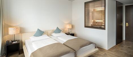 A hotel room with a double bed, two bedside tables with lamps, a wooden floor, and a window with blinds.