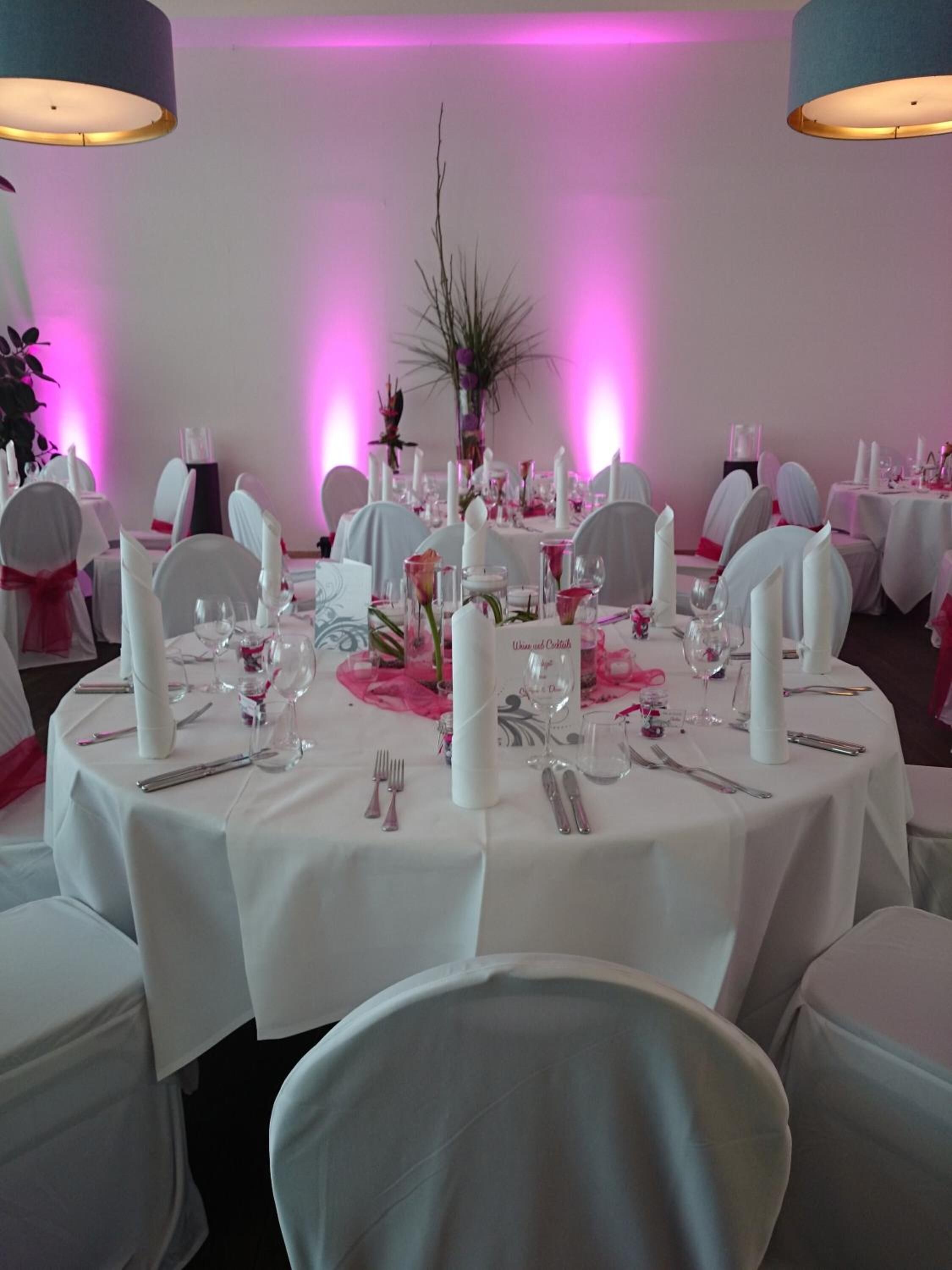 A round dining table set for a formal event with white tablecloths, silverware, and pink accents.