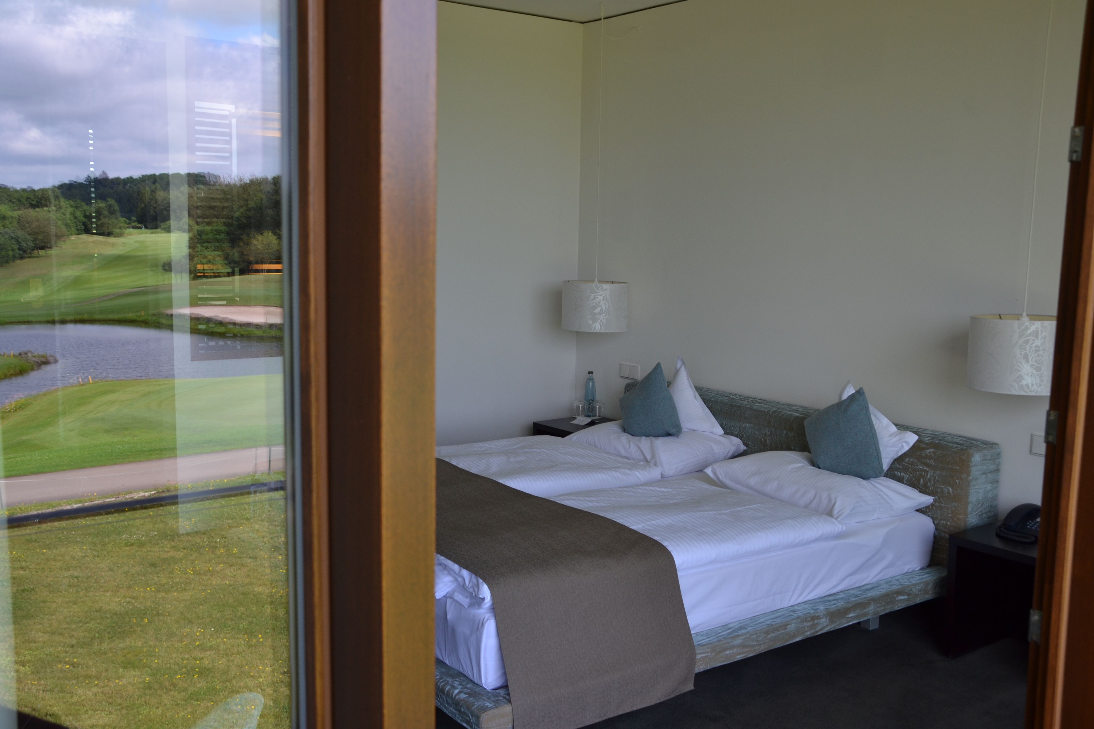 Habitación de hotel con un ventanal que ofrece vistas a un campo de golf y una masa de agua. La habitación tiene una cama con ropa blanca y dos almohadas, una mesita de noche con una lámpara y un pequeño mueble con un teléfono.