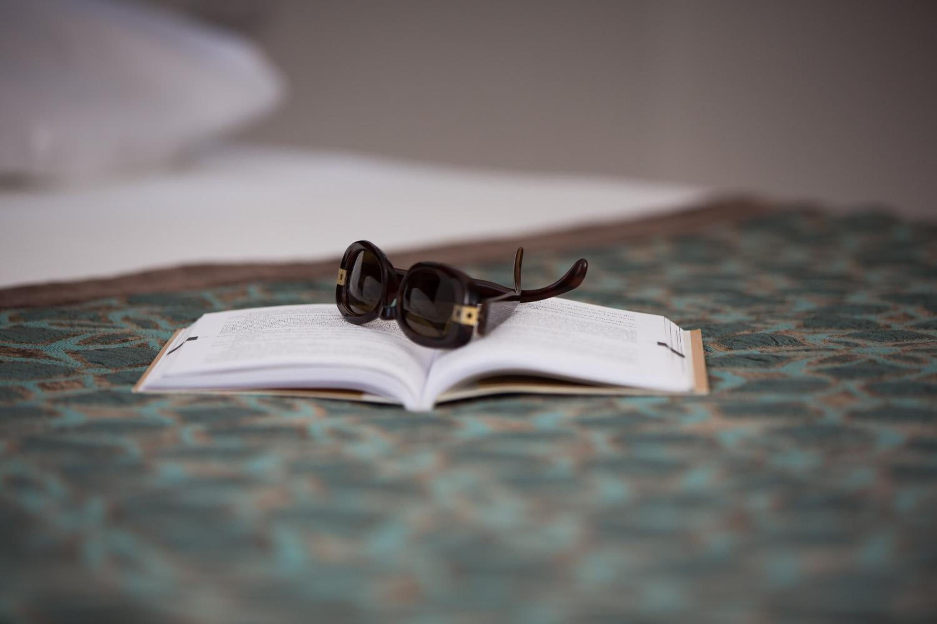 An open book with glasses on top, placed on a patterned fabric surface.