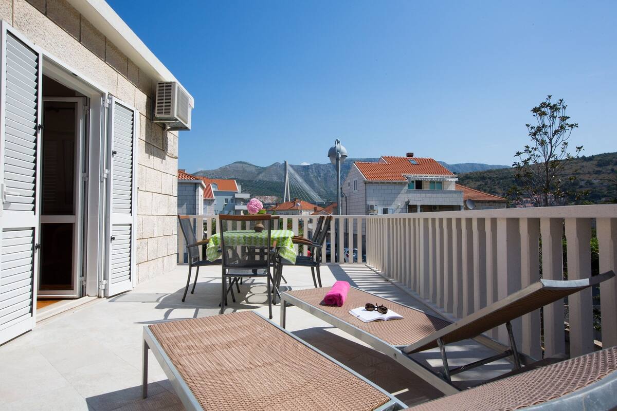 A balcony with a table and chairs, a view of mountains, and a building with a tiled roof.