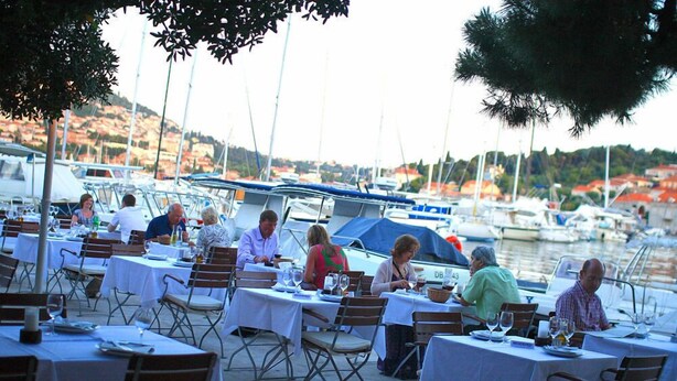 Un área de comedor al aire libre con mesas dispuestas para comidas, con vistas a un puerto deportivo con barcos y un paisaje montañoso al fondo.