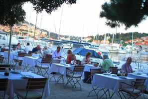 Un área de comedor al aire libre con mesas dispuestas para comidas, con vistas a un puerto deportivo con barcos y un paisaje montañoso al fondo.