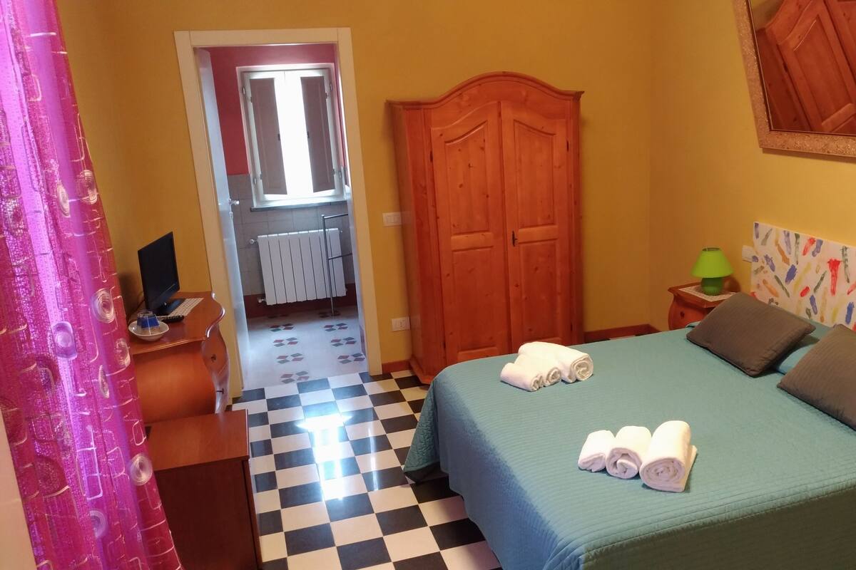 A bedroom with a checkered floor, a bed with pillows and towels, a wooden wardrobe, a desk with a computer, and a window with curtains.