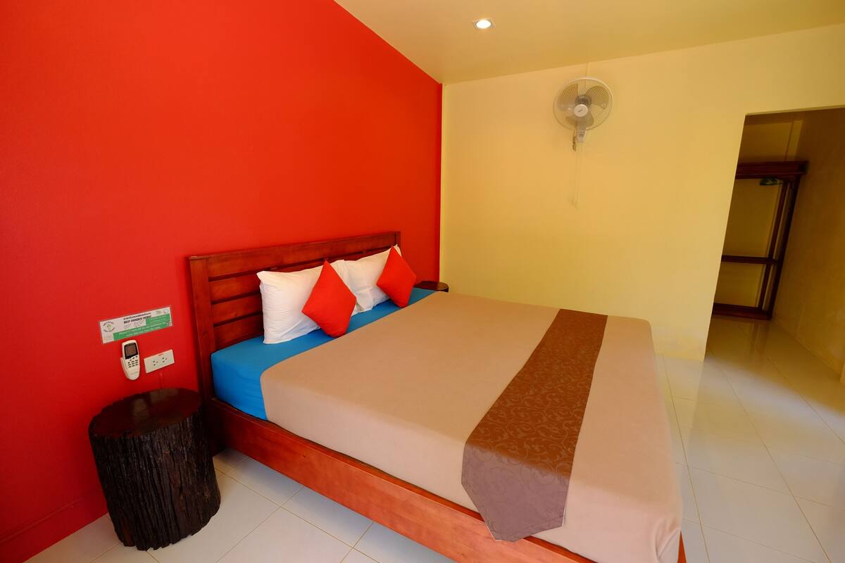 A hotel room with a wooden bed, red walls, a bedside table, and a fan on the wall.